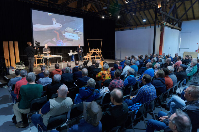 HolzWerken live in vollem Gange: Tischlermeister Guido Henn im Hintergrund auf der Bühne, viel Publikum auf Stühlen im Vordergrund