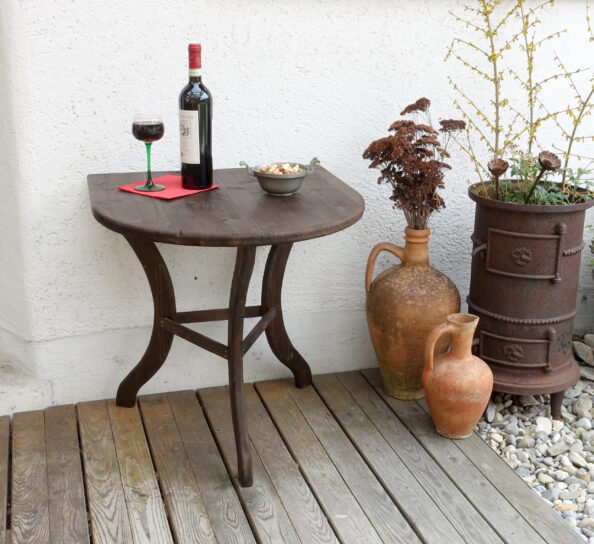 Auf einer Terrasse steht ein schokoladenbrauner halbrunder Beistelltisch mit geschwungenen Beinen, gedeckt mit Weinglas und Weinflasche. Daneben Amphoren mit Blumen darin und ein alter gußeiserner zylindrischer Ofen.