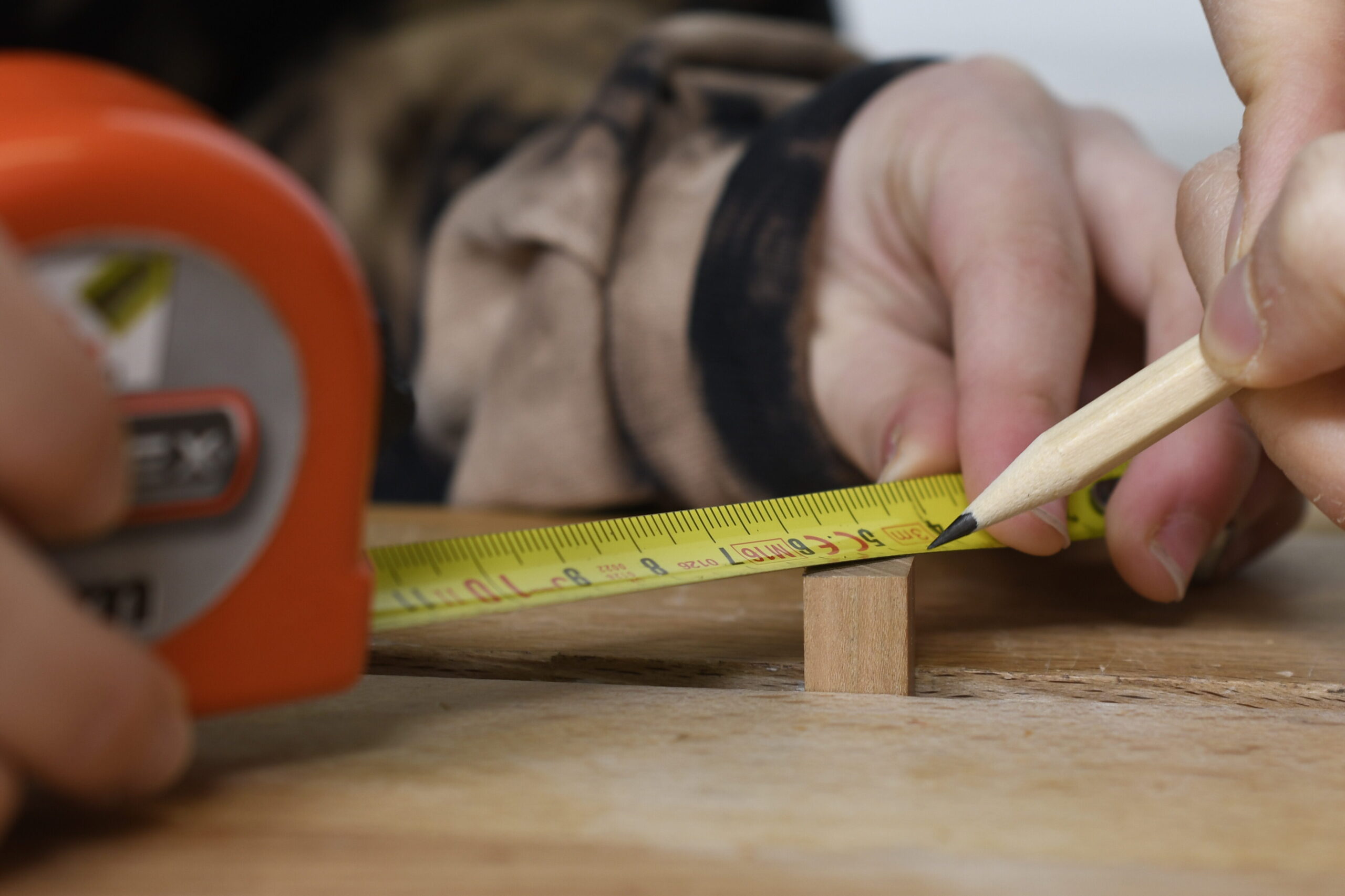 Die Hände von zwei Jugendlichen zeichnen den Mittelpunkt eines qadratischen Werkstücks an. Zwei Hände halten ein Bandmaß über die Diagonale, eine Hand hält einen spitzen Bleistift, um die Markierung zu setzen.