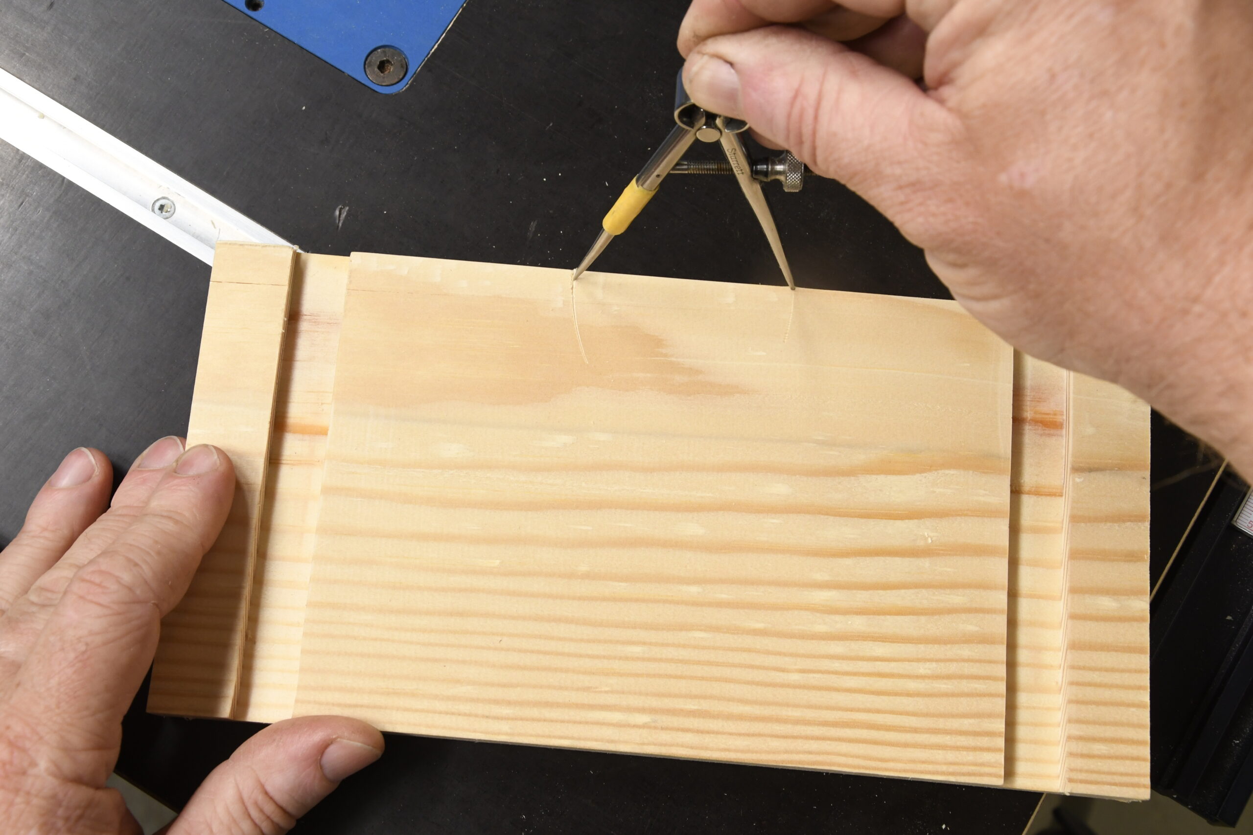 Ein Werkstück mit zwei breiten Nuten wird in die Kamera gehalten. Eine zweite Hand hält einen Zirkel und misst eine Strecke an der oberen Kante des Werkstückes ab.